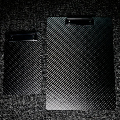 Close-up detail of two 3K carbon fiber clipboards featuring a distinctive woven pattern and black metal clips, positioned on a dark textured surface.