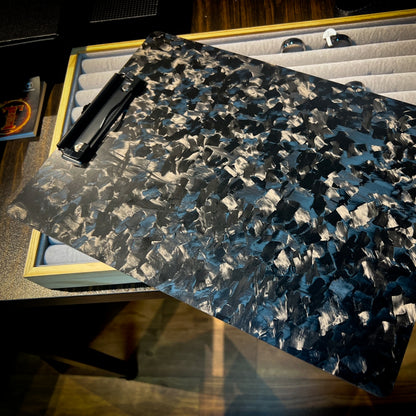 Close-up detail of a forged carbon fiber clipboard with a distinct marbled texture and a black metal clip, positioned over a display case with a wooden frame and gray padding.