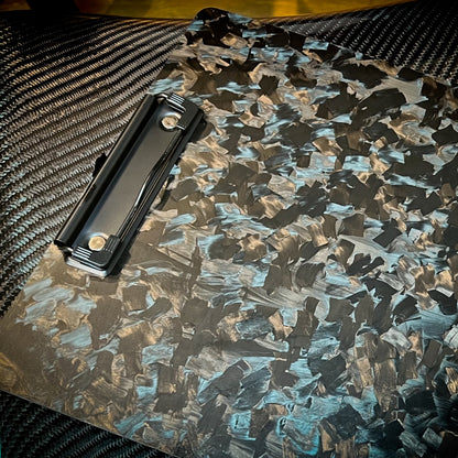 Close-up detail of a forged carbon fiber clipboard with a distinct marbled chopped pattern and a black metal clip, resting on a carbon fiber-textured surface.