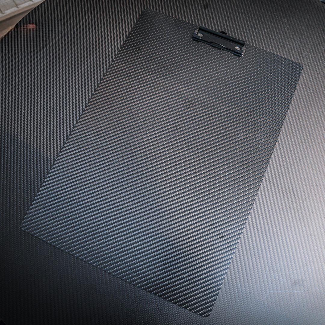 Close-up detail of a 3K carbon fiber clipboard showcasing its signature woven pattern, equipped with a black metal clip, placed on a carbon fiber-textured surface.