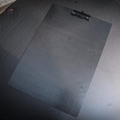 Close-up detail of a 3K carbon fiber clipboard showcasing its signature woven pattern, equipped with a black metal clip, placed on a carbon fiber-textured surface.