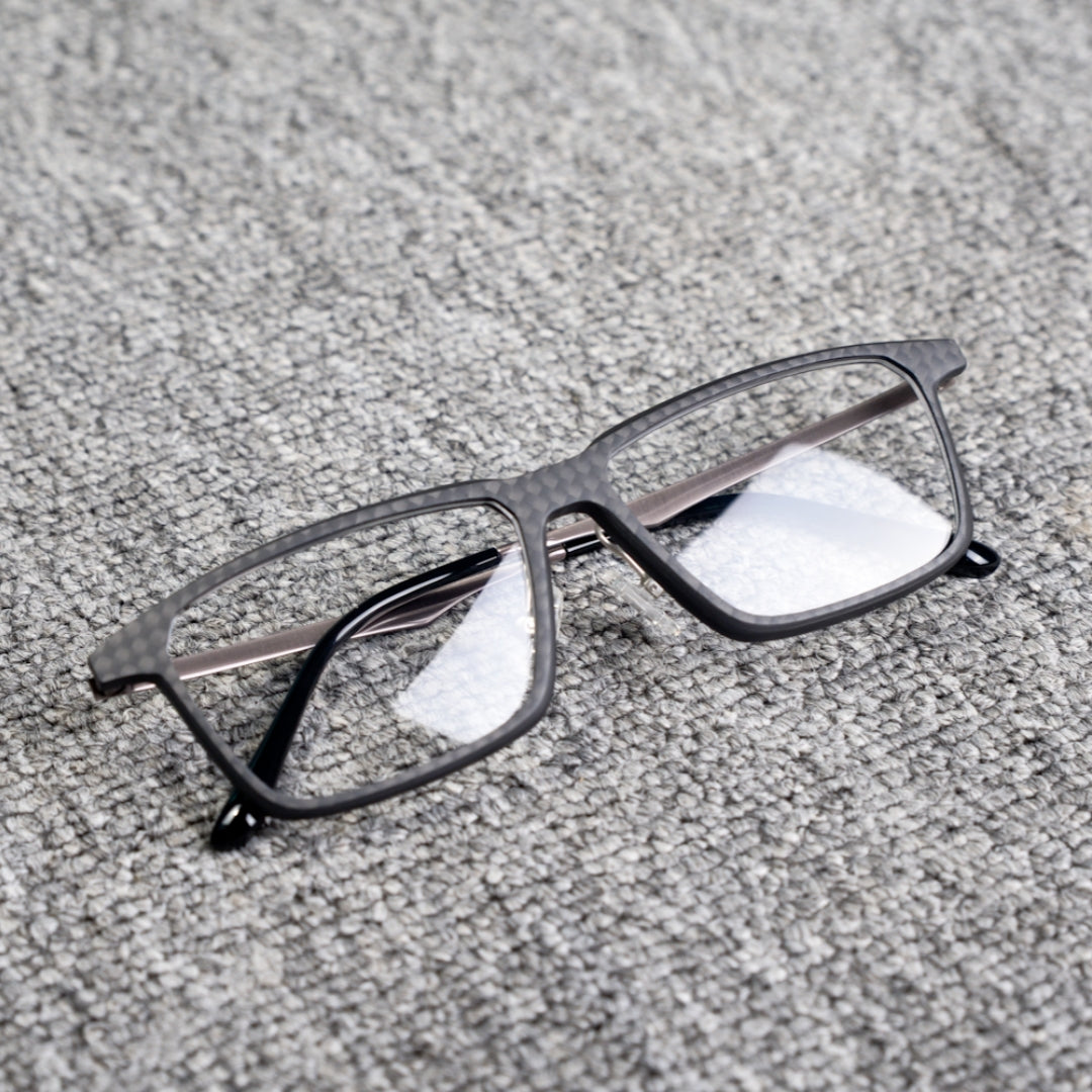 Close-up of a 3K plain weave carbon fiber eyeglass frame with a square shape and clear lenses, placed on a textured gray surface.
