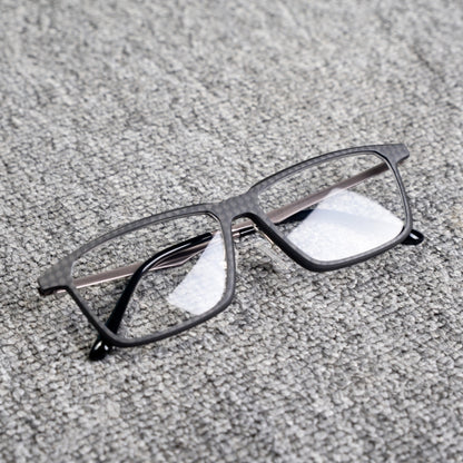 Close-up of a 3K plain weave carbon fiber eyeglass frame with a square shape and clear lenses, placed on a textured gray surface.
