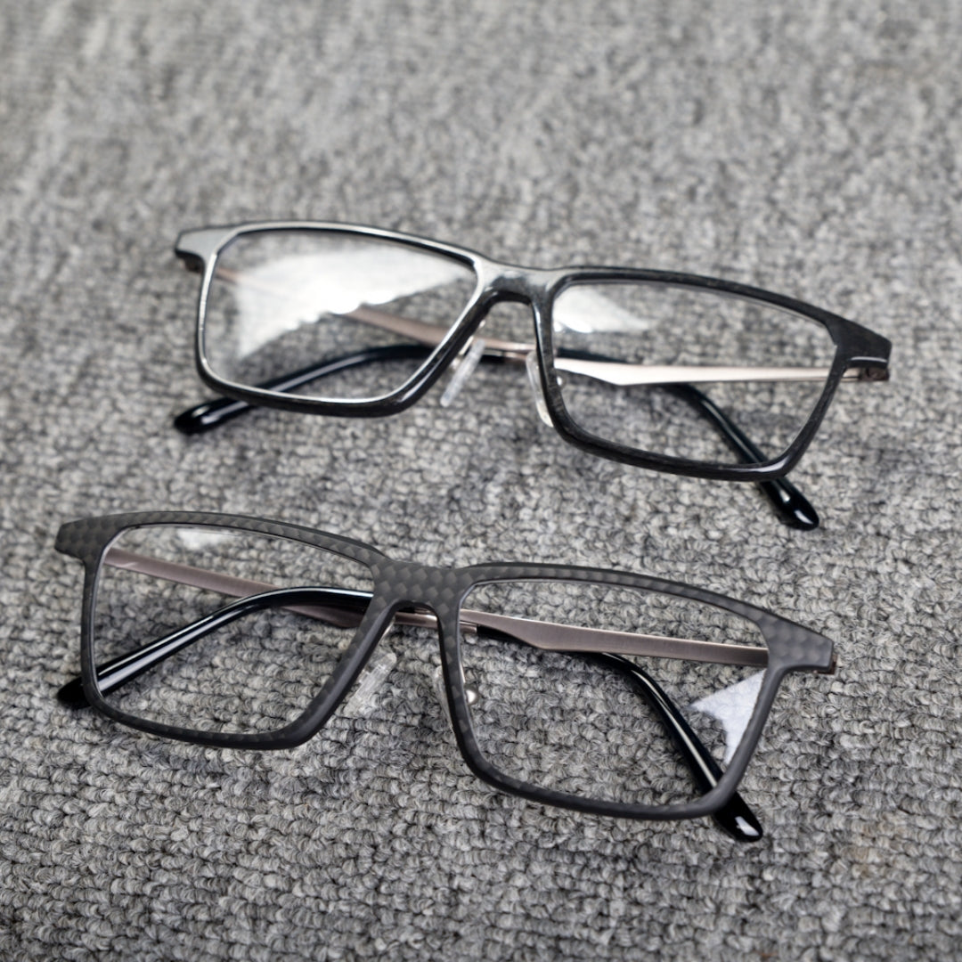 Close-up of two eyeglass frames featuring forged carbon fiber and 3K carbon fiber constructions, with sleek black designs and clear lenses, placed on a textured gray surface.