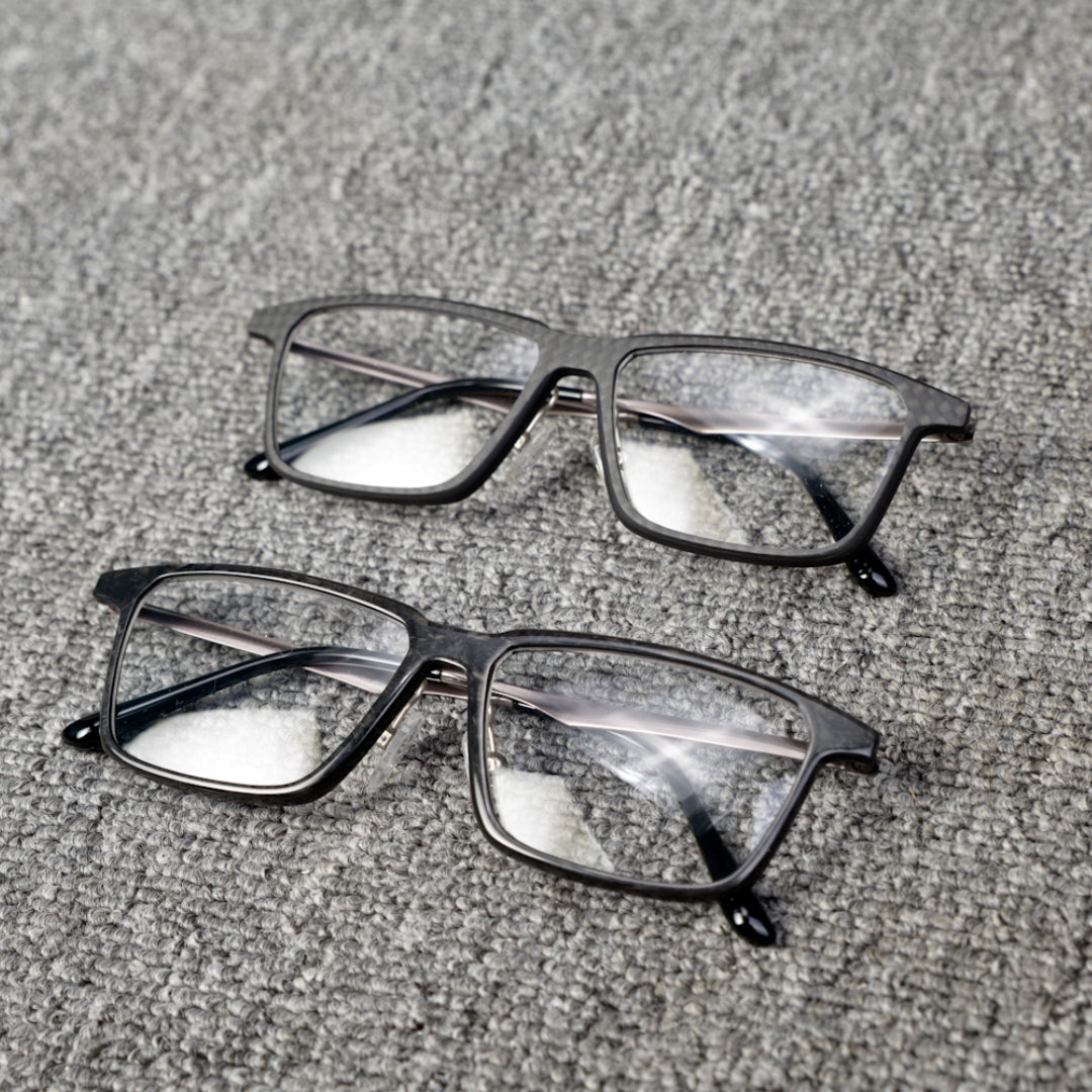 Close-up of two pairs of eyeglass frames crafted from forged carbon fiber and 3K carbon fiber, showcasing sleek black designs with clear lenses, placed on a textured gray surface.