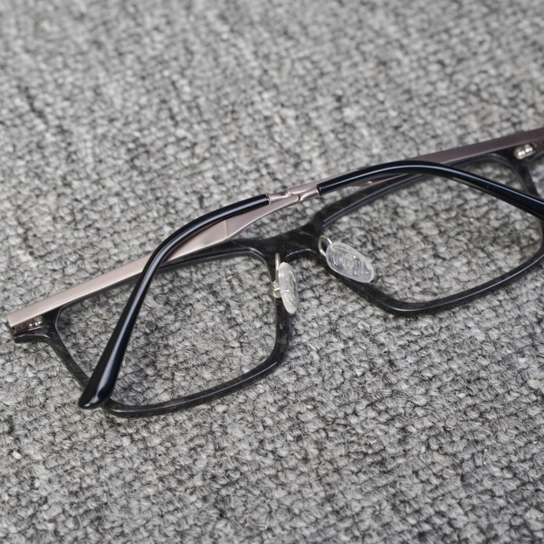 Close-up reverse side detail of a forged carbon fiber eyeglass frame, featuring clear nose pads and black with metallic temples, placed on a textured gray surface.