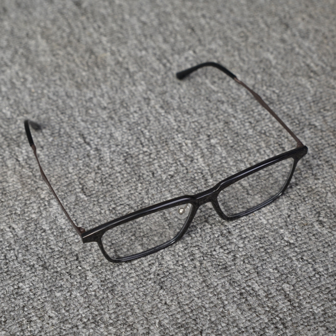 Close-up top detail of a forged carbon fiber eyeglass frame with a sleek black rectangular design, placed on a textured gray surface.