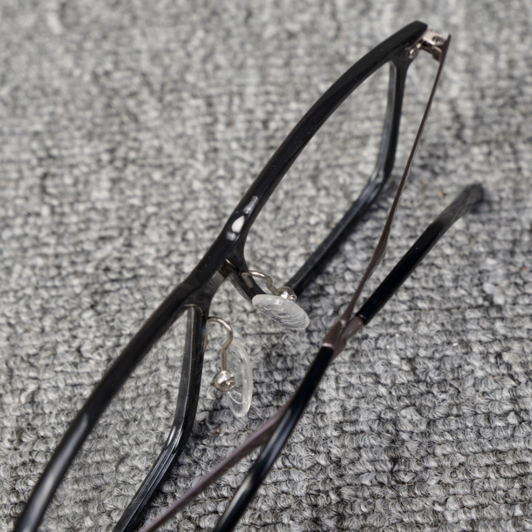 Close-up top detail of a forged carbon fiber eyeglass frame with a sleek black design, clear nose pads, and metallic components, placed on a textured gray surface.