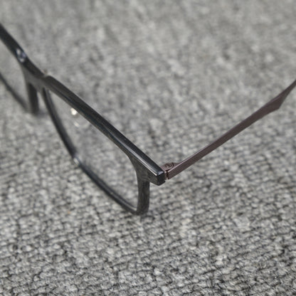 Close-up side detail of a forged carbon fiber eyeglass frame, showcasing the sleek black frame and metallic temple connection, placed on a textured gray surface.