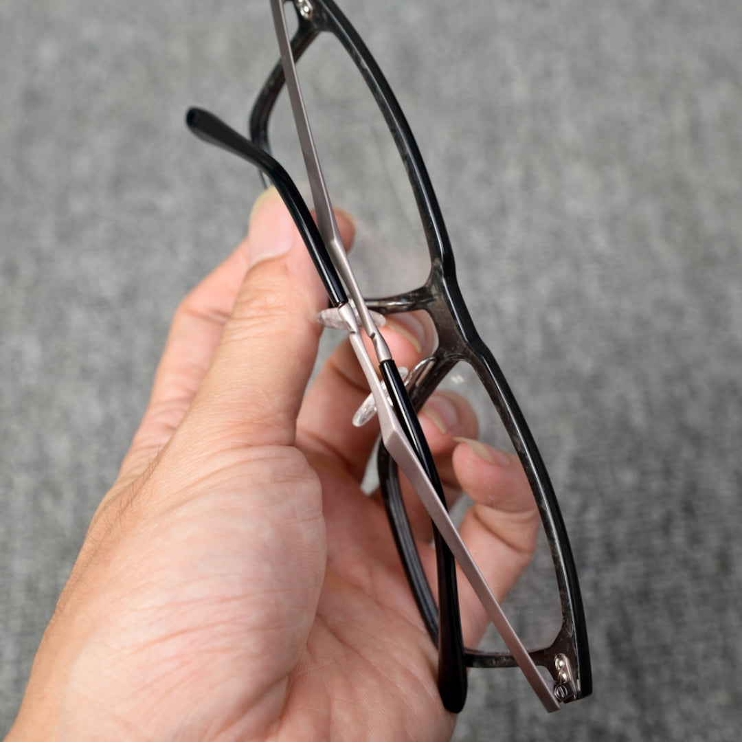 Close-up reverse side detail of a hand-held forged carbon fiber eyeglass frame, showcasing the sleek black frame and metallic components, against a textured gray background.