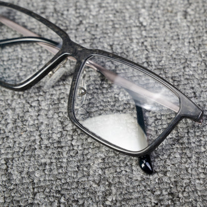 Close-up detail of a forged carbon fiber eyeglass frame with a sleek black design and clear lenses, placed on a textured gray surface.