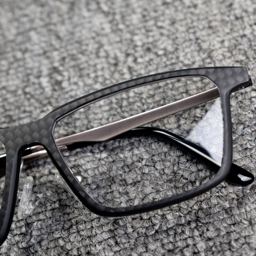 Close-up detail of a 3K carbon fiber eyeglass frame with a sleek black textured design and metallic temples, placed on a textured gray surface.
