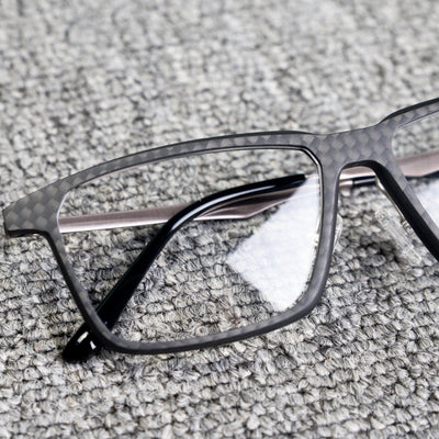 Close-up detail of a 3K carbon fiber eyeglass frame featuring a sleek black textured pattern, metallic temples, and clear lenses