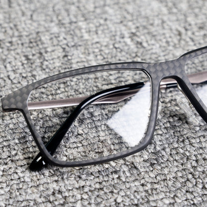 Close-up detail of a 3K carbon fiber eyeglass frame featuring a sleek black textured pattern, metallic temples, and clear lenses, placed on a textured gray surface.