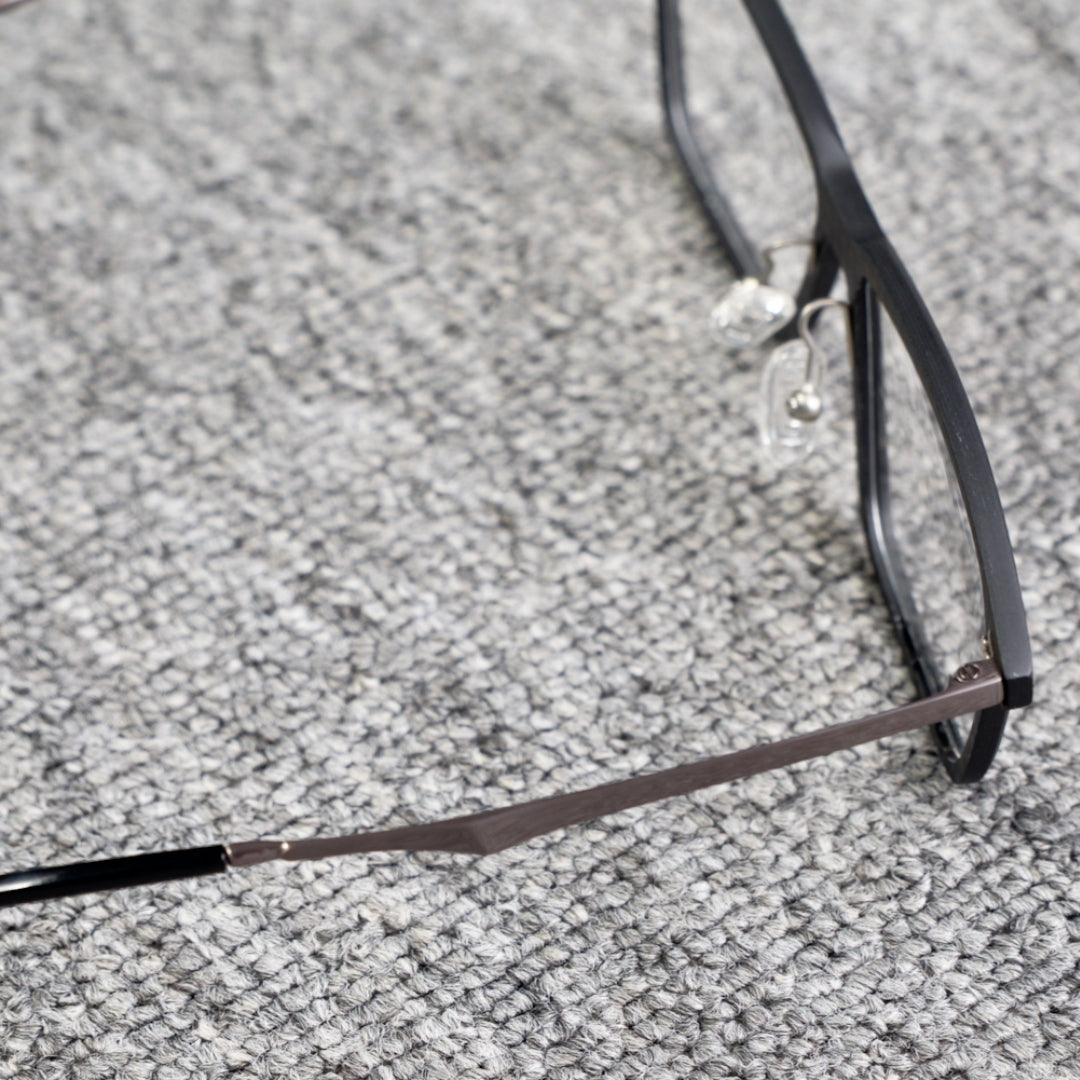 Close-up side detail of a 3K carbon fiber eyeglass frame, featuring a sleek black design and metallic temple, placed on a textured gray surface.