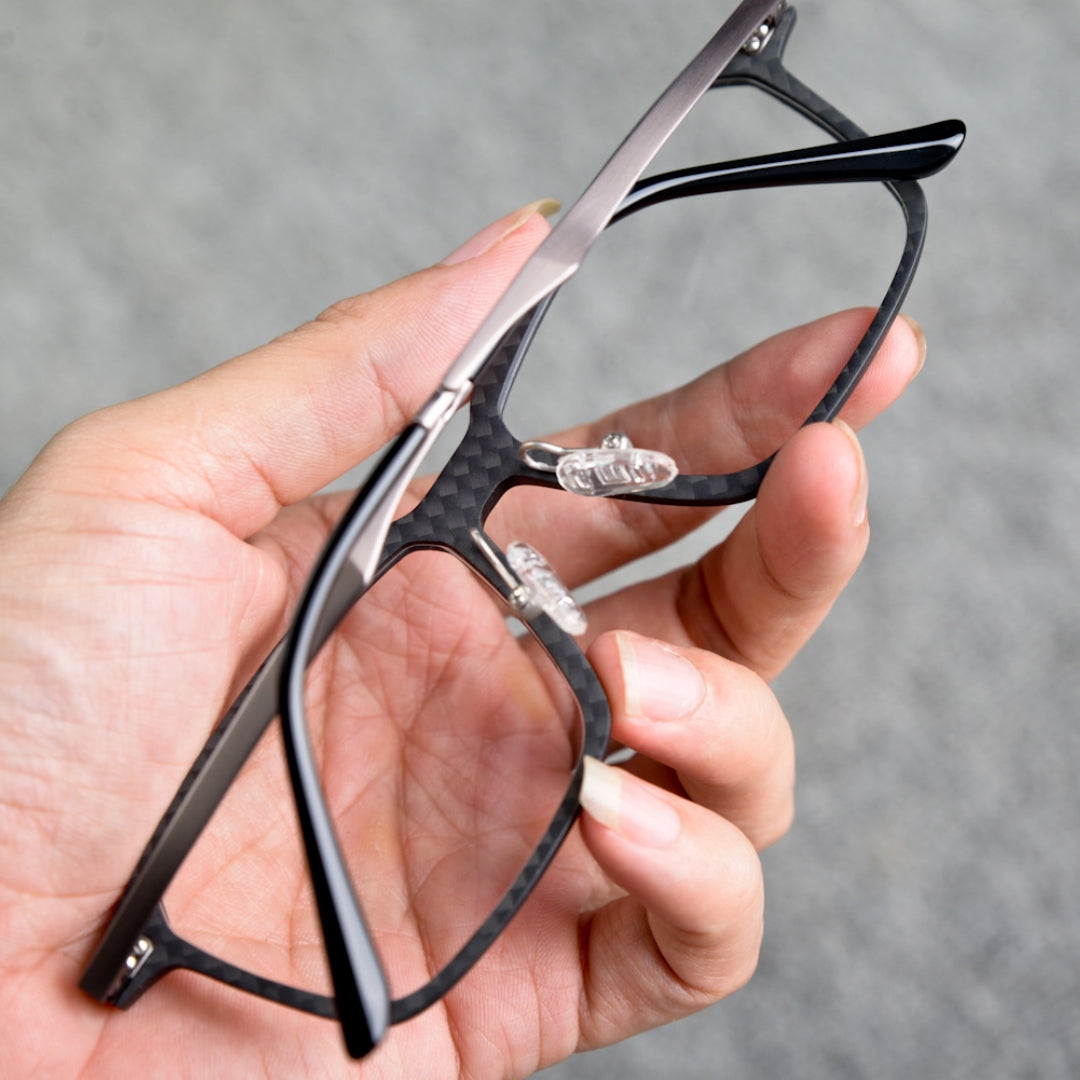 Close-up reverse side detail of a hand-held 3K carbon fiber eyeglass frame, showcasing its sleek black carbon fiber pattern, clear nose pads, and metallic/black temples, against a neutral gray background.