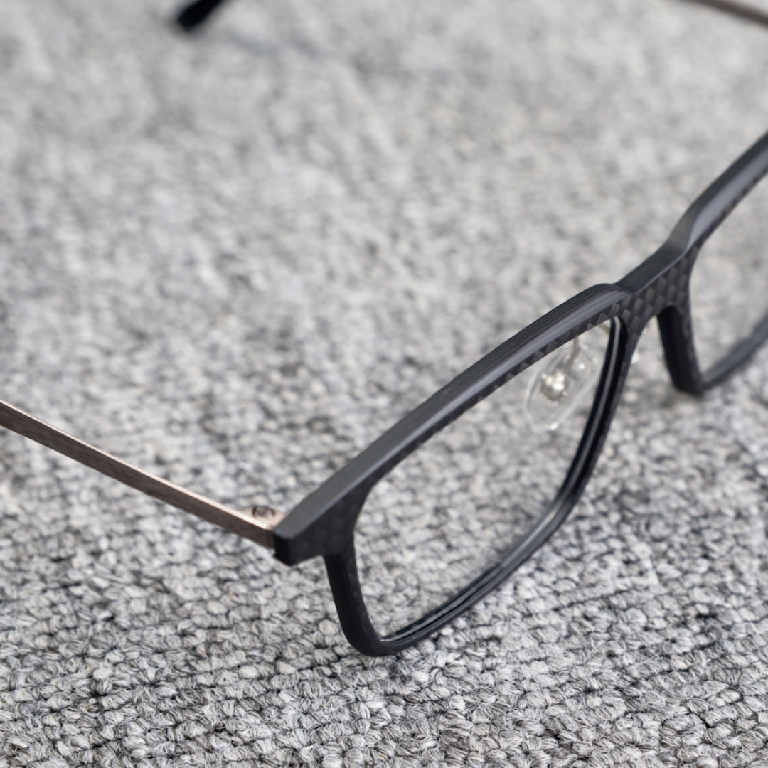 Close-up side detail of a 3K carbon fiber eyeglass frame, highlighting its sleek black carbon fiber pattern and metallic temple, placed on a textured gray surface.