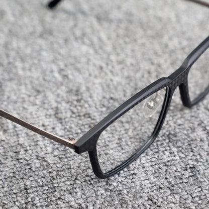 Close-up side detail of a 3K carbon fiber eyeglass frame, highlighting its sleek black carbon fiber pattern and metallic temple, placed on a textured gray surface.