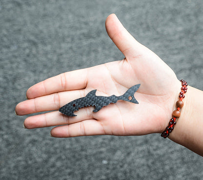 Close-up of a shark-shaped 3K carbon fiber keychain with a checkered pattern and serrated edge details, resting on an open palm adorned with a red and brown beaded bracelet, against a gray textured background.