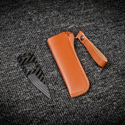 Close-up of a carbon fiber knife with a striped blade and a carbon fiber-patterned handle, paired with a brown leather sheath that has a key ring, displayed on a dark gray textured fabric surface.