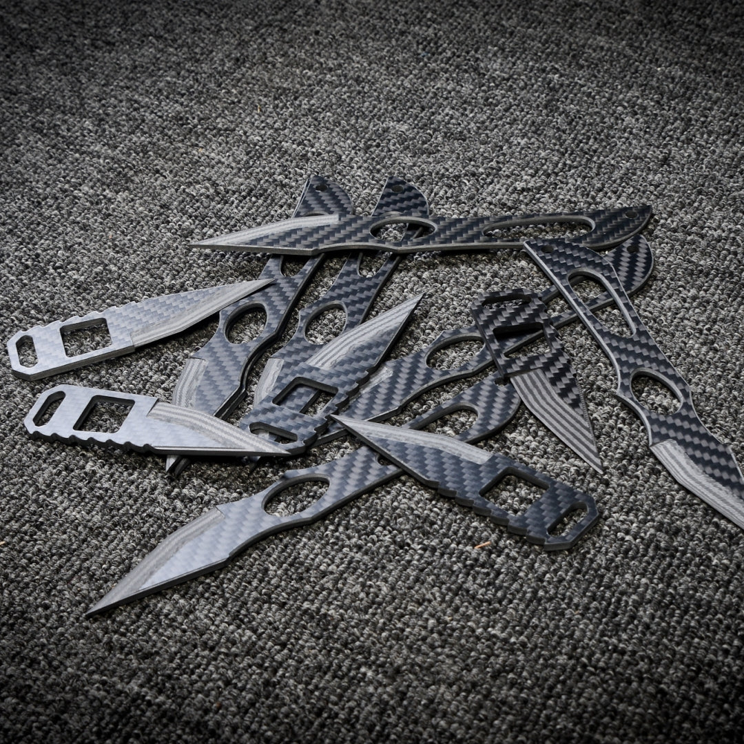 Close-up of multiple carbon fiber knives with various designs, showcasing their sleek carbon fiber patterns, arranged on a dark gray textured fabric surface.