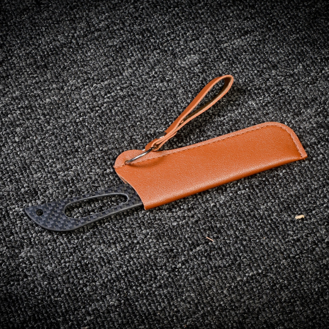 Close-up of a carbon fiber knife with a textured handle, sheathed in a brown leather case featuring a strap and key ring, placed on a dark textured fabric surface.