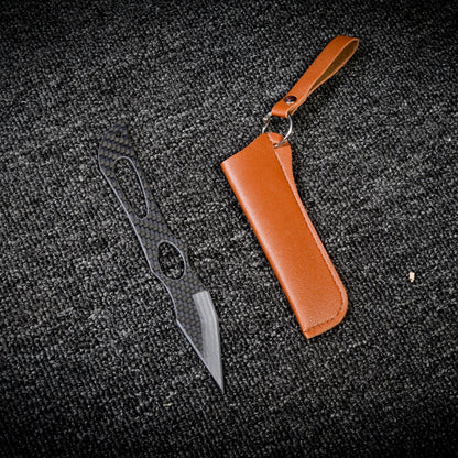 Close-up of a carbon fiber tactical knife with a patterned handle and blade, paired with a brown leather sheath featuring a key ring and strap, displayed on a dark textured fabric surface.