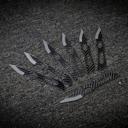 Close-up of multiple carbon fiber throwing knives with distinctive patterned handles and blades, arranged on a dark textured fabric surface.