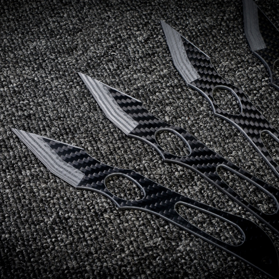 Close-up of multiple carbon fiber throwing knives with a checkered pattern and cutout designs, laid out on a dark textured fabric surface.
