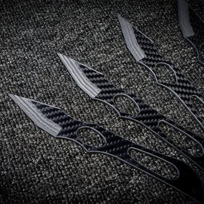 Close-up of multiple carbon fiber throwing knives with a checkered pattern and cutout designs, laid out on a dark textured fabric surface.