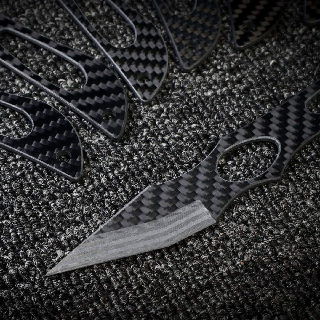 Close-up of multiple carbon fiber knives with a checkered pattern and cutout details, with one prominent knife showcasing a sharp blade, arranged on a dark textured fabric surface.