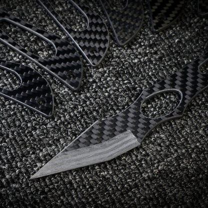 Close-up of multiple carbon fiber knives with a checkered pattern and cutout details, with one prominent knife showcasing a sharp blade, arranged on a dark textured fabric surface.