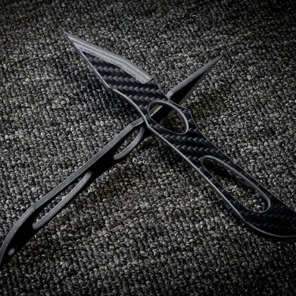 Close-up of two carbon fiber knives with intricate cutout designs and a checkered pattern, crossed over each other on a dark textured fabric surface.