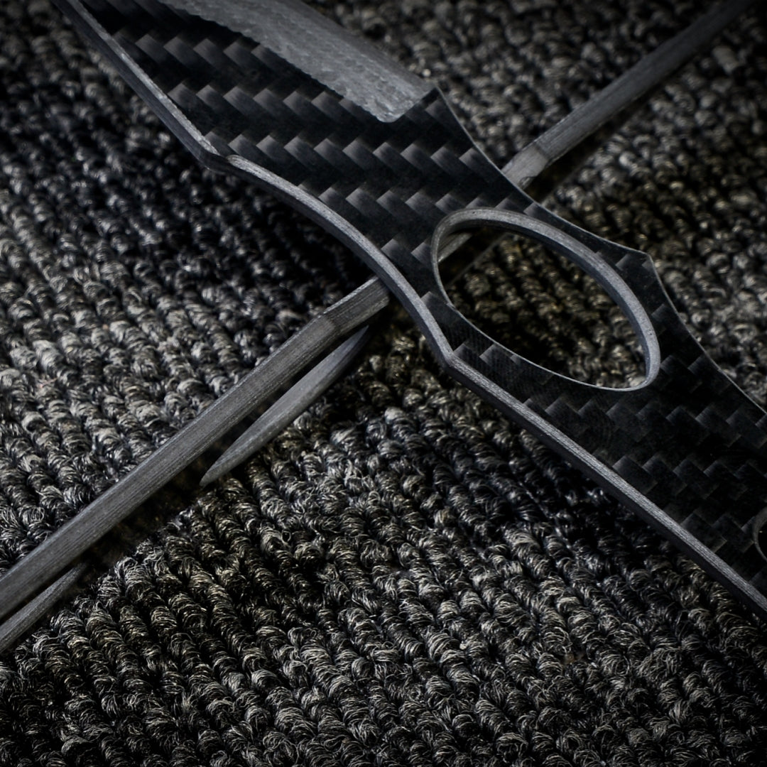 Close-up of two 3K carbon fiber knives with checkered patterns and cutout details, crossed over each other on a dark textured fabric surface.