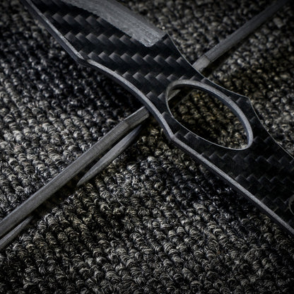 Close-up of two 3K carbon fiber knives with checkered patterns and cutout details, crossed over each other on a dark textured fabric surface.