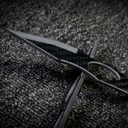 Close-up of two 3K carbon fiber knives with checkered patterns and cutout details, crossed over each other on a dark textured fabric surface.