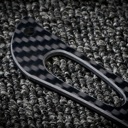 Close-up of a 3K carbon fiber knife handle with a checkered pattern and cutout detail, placed on a dark textured fabric surface.