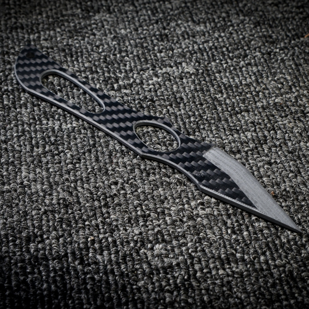 Close-up of a 3K carbon fiber knife with a checkered pattern and multiple cutout details on the handle, showcasing a sleek design with a sharp, metallic blade, placed on a dark textured fabric surface.