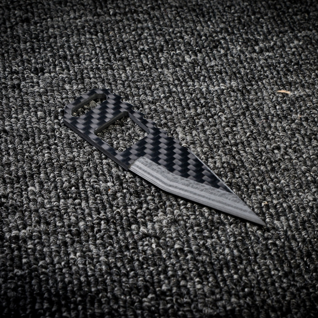 Close-up of a 3K carbon fiber knife featuring a checkered pattern, cutout handle design, and a sharp blade with metallic detailing, placed on a dark textured fabric surface.
