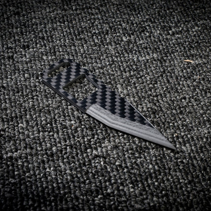 Close-up of a 3K carbon fiber knife featuring a checkered pattern, cutout handle design, and a sharp blade with metallic detailing, placed on a dark textured fabric surface.
