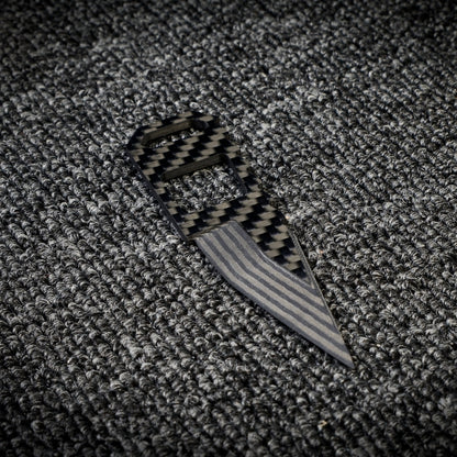 Close-up of a 3K carbon fiber knife with a checkered pattern handle and a striped metallic blade, placed on a dark textured fabric surface.