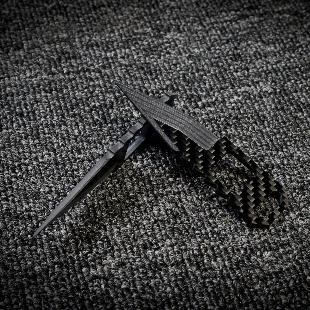 Close-up of two 3K carbon fiber knives with checkered patterns and cutout details, crossed over each other on a dark textured fabric surface.