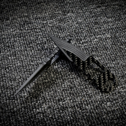 Close-up of two 3K carbon fiber knives with checkered patterns and cutout details, crossed over each other on a dark textured fabric surface.