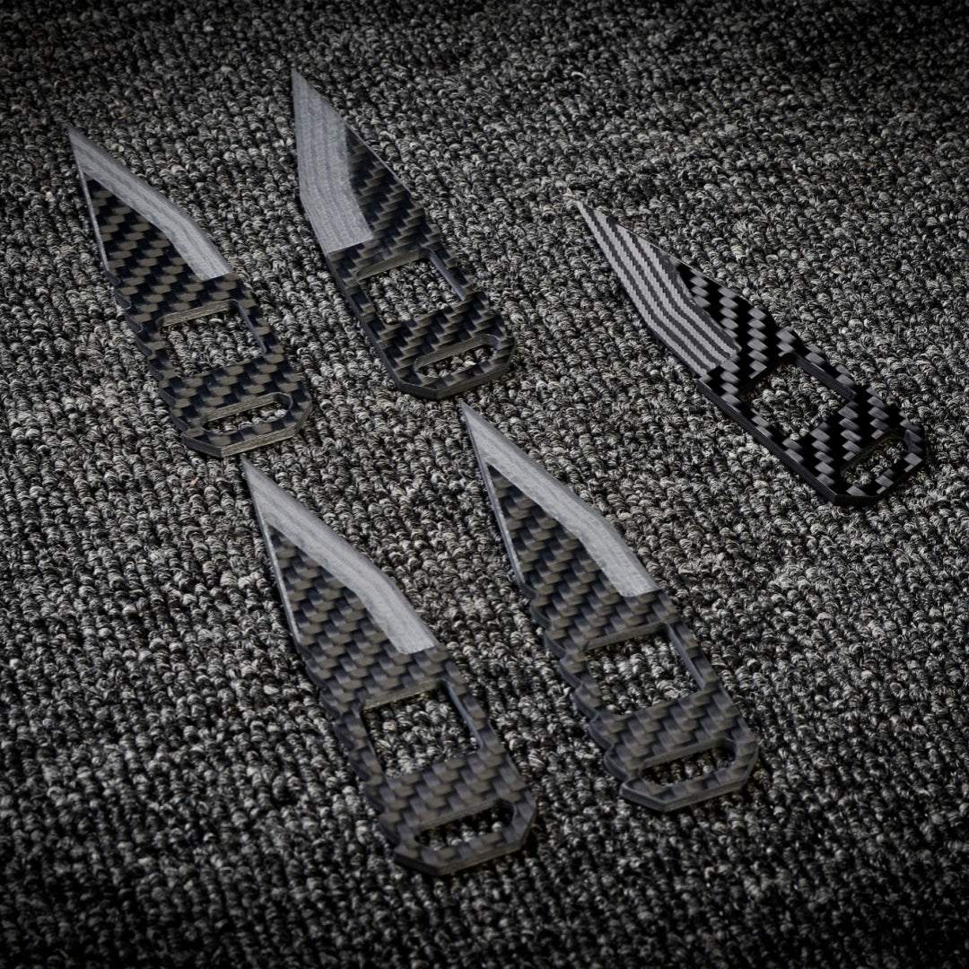 Close-up of five 3K carbon fiber knives with checkered patterns and varied cutout designs, arranged on a dark textured fabric surface.