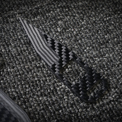 Close-up of a 3K carbon fiber knife featuring a checkered pattern, striped blade, and cutout details on the handle, positioned on a dark textured fabric surface.