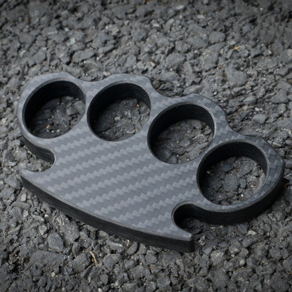 Close-up of a carbon fiber brass knuckles with four finger holes, showcasing its distinct woven pattern, placed on a gravel surface.