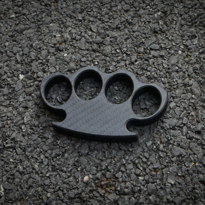 Close-up of a carbon fiber brass knuckles with four finger holes, showcasing its distinct woven pattern, placed on a gravel surface.