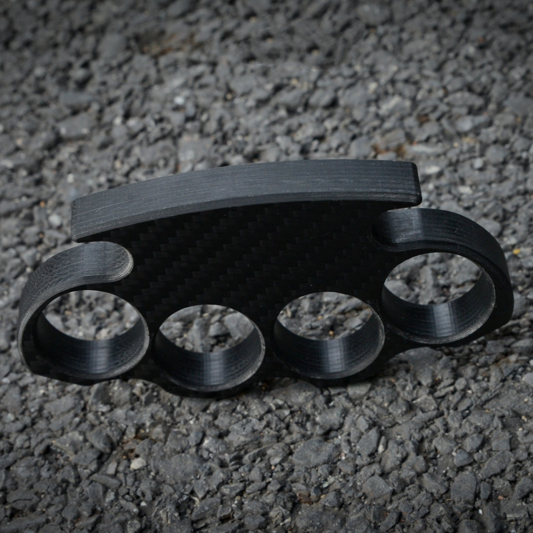 Close-up of a carbon fiber brass knuckles with four finger holes, displaying its textured carbon fiber pattern, placed on a gravel surface.