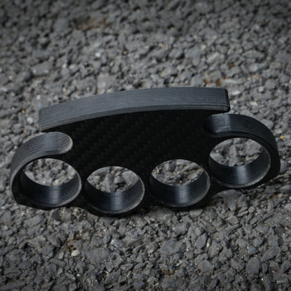 Close-up of a carbon fiber brass knuckles with four finger holes, displaying its textured carbon fiber pattern, placed on a gravel surface.