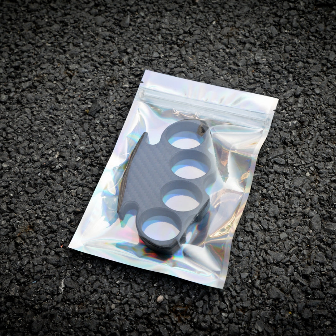 Close-up of a carbon fiber brass knuckles with four finger holes, enclosed in a holographic zip-lock bag, placed on an asphalt surface.